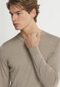 Beige långärmad skjorta gjord av mjukt tyg, med rund halsringning och ribbade ärmslut. Texturen verkar slät och lätt.