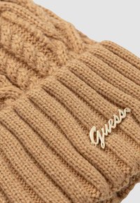 Berretto lavorato a maglia color cuoio con motivi a trecce e coste. Presenta un dettaglio con il logo "Guess" in tono oro sul risvolto. Texture morbida.