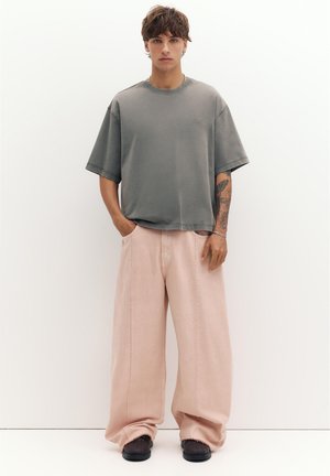 Jeune homme avec un bras tatoué portant un T-shirt gris ample, un pantalon large rose pâle, des chaussures noires, debout devant un fond blanc avec les mains partiellement dans les poches.
