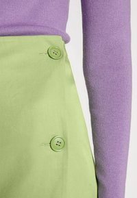 Saia de tecido verde com dois botões redondos verdes, combinada com um suéter roxo justo, exibindo textura canelada nas mangas.