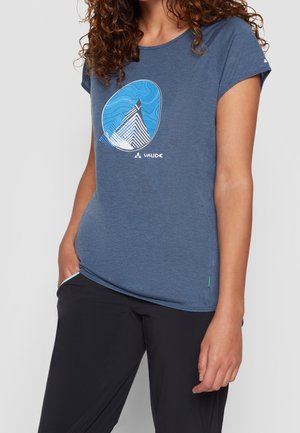 Vrouw draagt een blauw kortermig T-shirt met een blauw-wit berggrafiek en zwarte broek, staand met één hand in de zak.