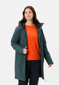 Donkergroene, waterdichte, knielange jas met een zwart gevoerde capuchon en een voorrits, gedragen over een oranje shirt. Beschikt over een verticale ritszak.