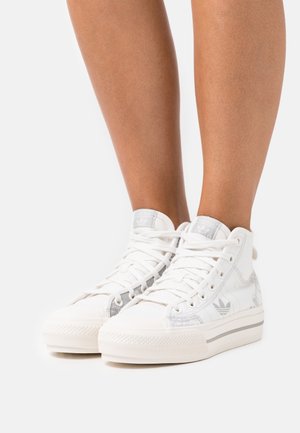 Nohy oblečené v bílých vysokých teniskách Adidas s jemnými šedými vzory a bílými tkaničkami, stojící na bílém pozadí.