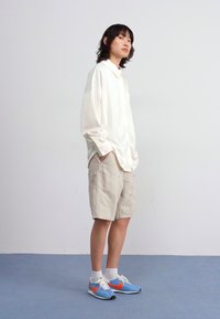 Vit långärmad skjorta, ljusbeige shorts och blå sneakers med orange detaljer. Slitstyg, avslappnad passform, står mot en enfärgad bakgrund.