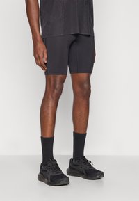 Μαύρο σορτσάκι από υλικό που απομακρύνει την υγρασία με εφαρμοστή σχεδίαση, διαθέτει επίπεδες ραφές. Συνδυάζεται με μαύρα αθλητικά παπούτσια και καλτσάκια. Απλό φόντο.