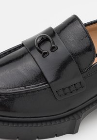 Black leather loafer with a textured finish, featuring a black strap and a decorative emblem. The sole is chunky and rubberized.