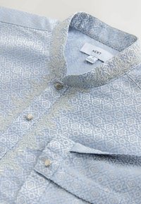 Hellblaues Hemd mit komplexen silbernen geometrischen Mustern, einem Stehkragen und Knopfverschluss. Weicher Stoff mit detaillierter Stickerei.