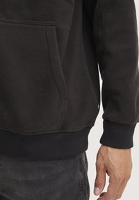Sudadera de forro polar negra con un bolsillo canguro, puños y dobladillo acanalados. La textura suave y el diseño sin costuras ofrecen un ajuste cómodo.