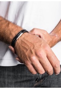 Brățară din piele împletită neagră, cu un închizător și accente metalice, odihnindu-se pe o mână. Tonul pielii este vizibil, evidențiind textura și detaliile.