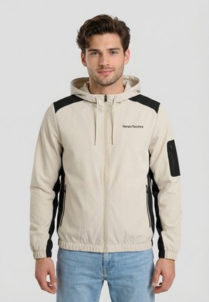 Joven con chaqueta beige con capucha y cremallera con detalles negros y vaqueros azul claro, de pie frente a un fondo claro y simple.