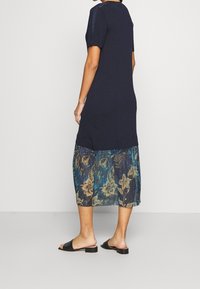 Granatowa sukienka z krótkim rękawem, z warstwową przezroczystą spódnicą w kwiatowe wzory w odcieniach niebieskiego i zielonego. Całość dopełniają czarne sandały.