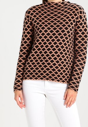 Woman wearing a long-sleeve black and peach diamond-patterned sweater with white pants, standing against a plain background.