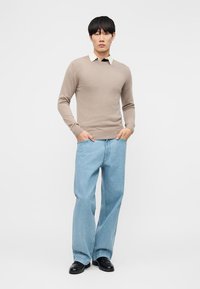 Homme portant un pull beige par-dessus une chemise blanche à col, un jean bleu clair à jambes larges, et des chaussures noires, debout les mains dans les poches.