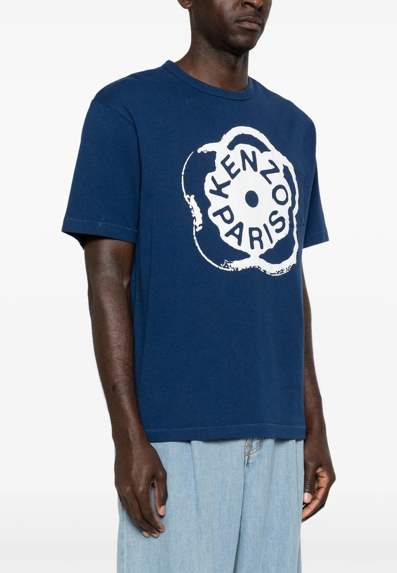 Mand iført en blå T-shirt med et hvidt rundt "Kenzo Paris" logo og lyseblå jeans, stående mod en ensfarvet hvid baggrund.