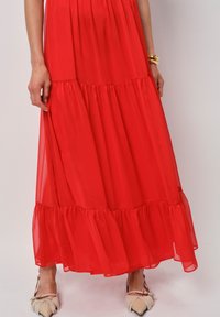 Rode maxi-jurk gemaakt van doorzichtig stof, met een gelaagd ontwerp en ruches aan de onderkant. Gecombineerd met neutrale platte schoenen.
