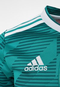 Vihreä adidas-pelipaita, jossa on kohokuvioitu raidallinen kuvio, valkoinen kaulus ja kolme valkoista raitaa olkapäillä. Logo valkoisena.