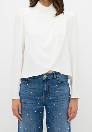 Kobieta w białej bluzce z długim rękawem i drapowanym przodem oraz dopasowanych, wysokim stanem niebieskich dżinsach ozdobionych małymi perłami.