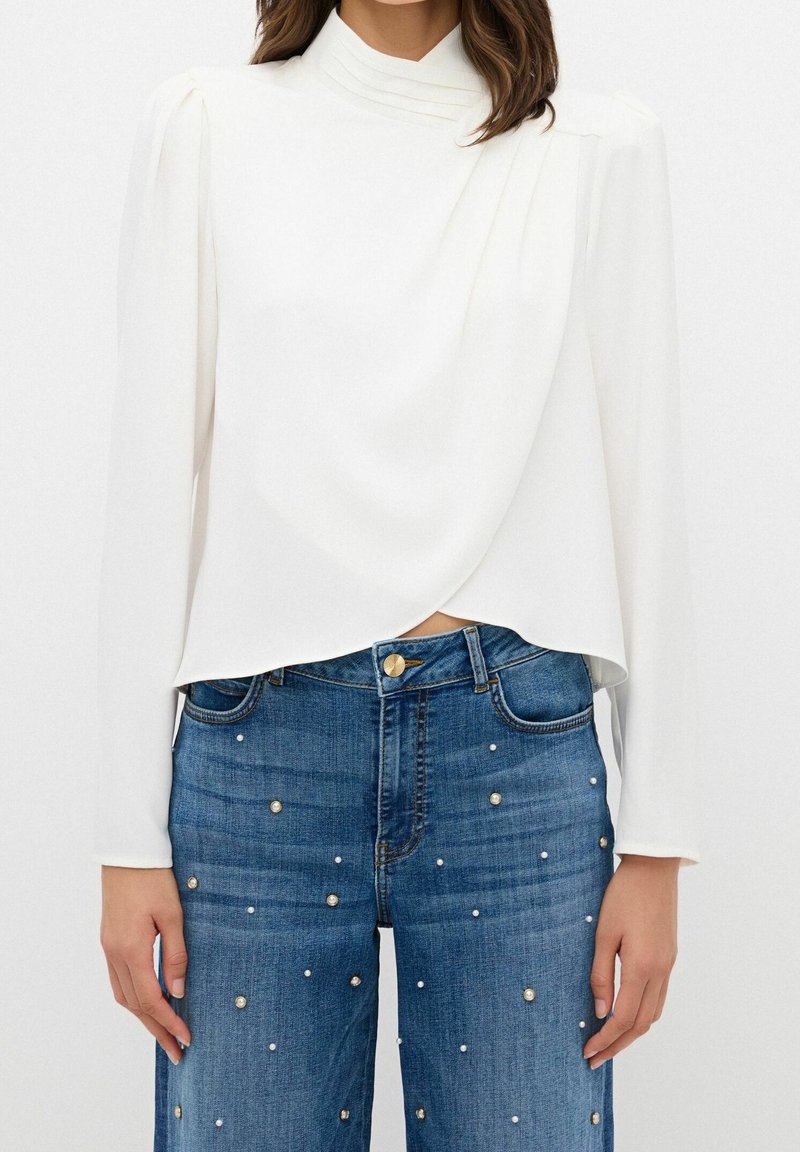 Femme portant un chemisier blanc à manches longues avec un devant drapé et un jean taille haute bleu orné de petites perles.