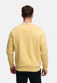 Gul bomullsgenser med lange ermer, rund hals, ribbede mansjetter og en smal avslutning nederst. Bak har den en buet skulderdetalj.