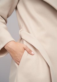 Beżowy, miękki płaszcz z tkaniny o luźnym kroju, z wiązanym paskiem i boczną kieszenią. Tekstura jest gładka i lekko puszysta.