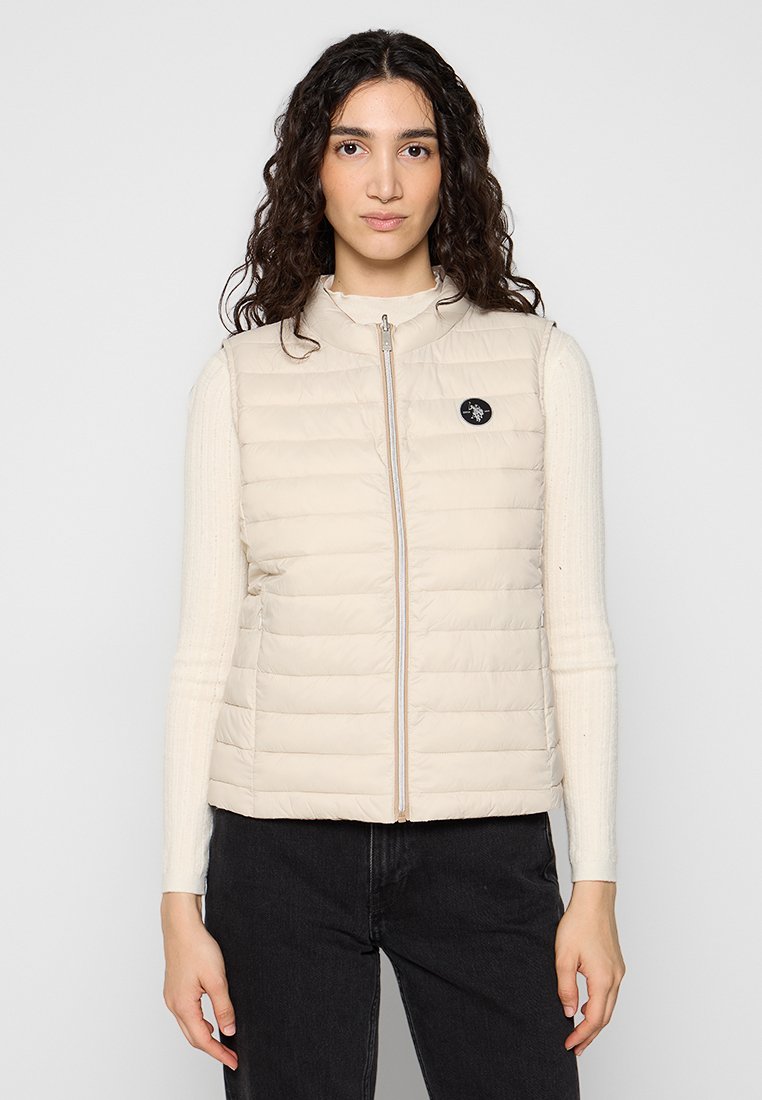 Eine Frau mit langen lockigen Haaren trägt eine beigefarbene gesteppten Weste über einem hellen Pullover und schwarzen Hosen und steht vor einem schlichten Hintergrund.