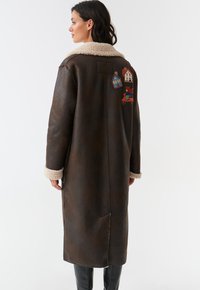 Manteau en cuir marron avec un col duveteux, orné de maisons brodées et d'un personnage au dos. Design long et droit avec des manches à revers.