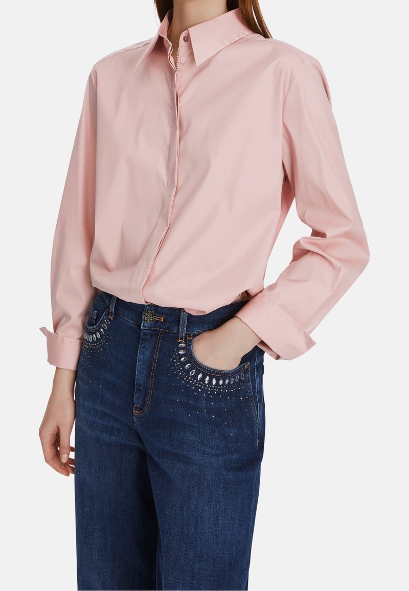 Chemise boutonnée rose clair à manches longues avec un col, portée rentrée dans un jean en denim bleu foncé orné de détails cloutés décoratifs.
