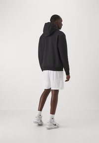 Sort hættebluse med en blød tekstur, parret med hvide mesh shorts. Modellen bærer hvide Nike sneakers med sorte accenter og sokker.