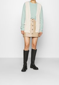 Débardeur côtelé beige clair avec poches porté sur un pull vert menthe, associé à une jupe boutonnée en cuir marron clair et des bottes noires montantes.