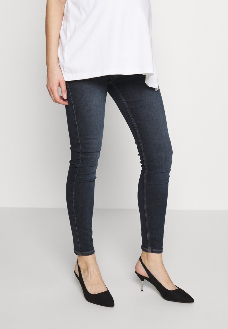 Mujer con jeans ajustados oscuros, tacones negros de tirantes y una camisa blanca oversize contra un fondo claro liso.