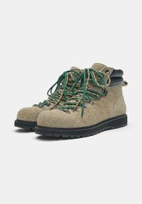 Para beżowych, teksturowanych butów trekkingowych z zielono-beżowym sznurowaniem, czarnymi, wyściełanymi kołnierzami i grubą czarną gumową podeszwą na białym tle.