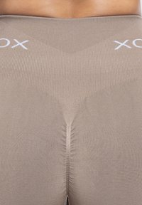 Beige naadloze leggings met een geribbelde textuur. Kenmerkt een V-vormige taille en een wit logo op de tailleband, wat het strakke ontwerp benadrukt.