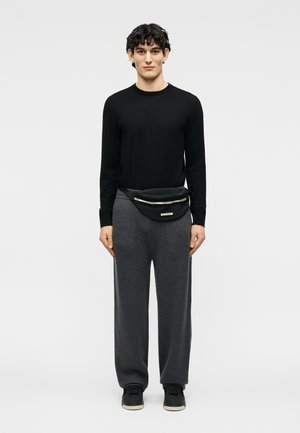 Jeune homme aux cheveux bouclés portant une chemise noire à manches longues, un pantalon gris, des baskets noires et une sacoche noire à glissières.