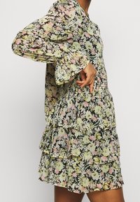 Abito floreale con maniche lunghe, volant sovrapposti e vita smock. Fiori giallo chiaro, rosa e verdi su uno sfondo nero. Tessuto trasparente.