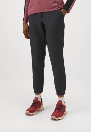 Personne portant un pantalon de jogging noir avec des poignets élastiques et des chaussures de randonnée rouges avec des chaussettes blanches, debout devant un fond uni.