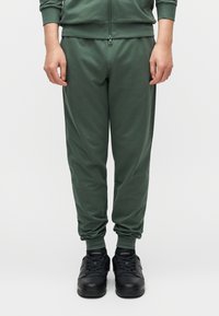 Πράσινο, στενό παντελόνι φόρμας κατασκευασμένο από μαλακό ύφασμα· διαθέτει ελαστικά μανσέτα και μέση· συνδυασμένο με μαύρα αθλητικά παπούτσια.