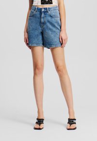 Denim kratke hlače v svetlo modri barvi z višjim pasom, sprednjimi žepi in neobdelanim robom. Nosi jih s črnimi ravnimi sandalmi. Gladka tekstura.