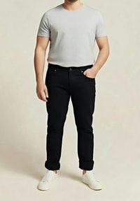 Homme portant une chemise à manches courtes gris clair, un pantalon noir retroussé et des baskets blanches, debout avec une main dans la poche.