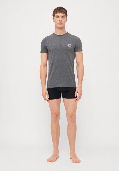 Graues T-Shirt mit kurzen Ärmeln und rundem Ausschnitt sowie kleinem Logo, kombiniert mit schwarzen, eng anliegenden Shorts, barfuß vor einem weißen Hintergrund stehend.