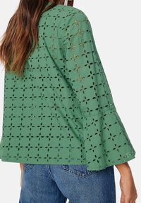 Haut vert et léger avec un motif floral en guipure, manches évasées et tissu texturé. Vue de dos avec détails de coutures visibles.