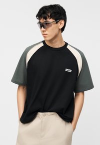 Ung mann med solbriller, iført en svart og grønn kortermet skjorte med teksten "kaotiko society" og beige bukser med hendene i lommene.