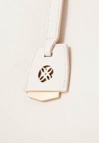 White textured handbag close-up with a gold accent and an intricate cutout logo on a leather tag. Features a smooth and sleek surface.