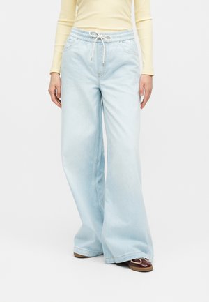 Osoba nosiaca svetlomodré široké džínsy so šnúrkou v páse v kombinácii so žltým dlhým rukávom a hnedými topánkami.