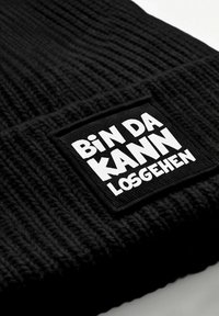 Black knitted fabric with a white patch displaying the German phrase "Bin da kann losgehen" in bold letters.