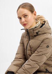 Bruine donzen jas met zwarte ritssluiting en capuchon, met een doorgestikte textuur en een hoge kraag. Voorzien van een cirkellogo op de mouw.