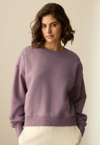 Ljubičasta oversize majica dugih rukava s okruglim izrezom i rebrastim manžetnama. Izrađena od mekanog, glatkog tkanja i ima opušteni kroj. Kombinirana s hlačama svijetle boje.