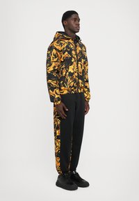 Man i stående pose iförd svart och guld barockmönstrad huva-jacka och matchande svarta byxor med mönstrade ränder och svarta sneakers.