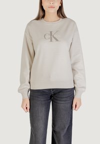 Sudadera beige claro con cuello redondo, puños y dobladillo acanalados. Presenta un logotipo gris de "Calvin Klein" impreso en la parte frontal. Jeans de mezclilla oscuros visibles.