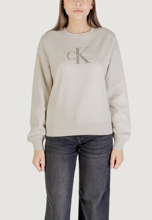 Sudadera beige claro con cuello redondo, puños y dobladillo acanalados. Presenta un logotipo gris de "Calvin Klein" impreso en la parte frontal. Jeans de mezclilla oscuros visibles.