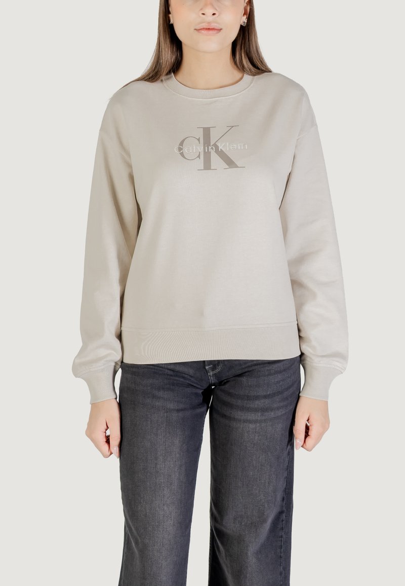 Sudadera beige claro con cuello redondo, puños y dobladillo acanalados. Presenta un logotipo gris de "Calvin Klein" impreso en la parte frontal. Jeans de mezclilla oscuros visibles.